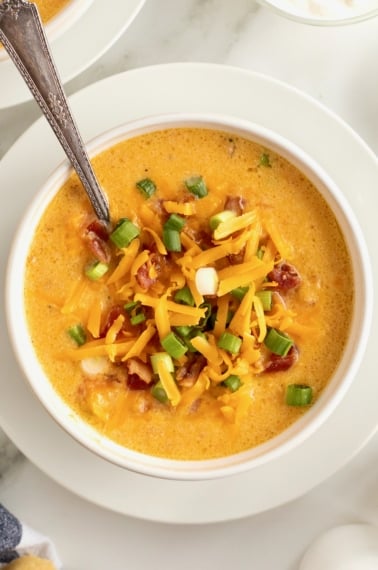 A white bowl of sweet potato soup on a white saucer with a spoon in it. The soup is topped with bacon bits, shredded yellow cheese and sliced green onion.