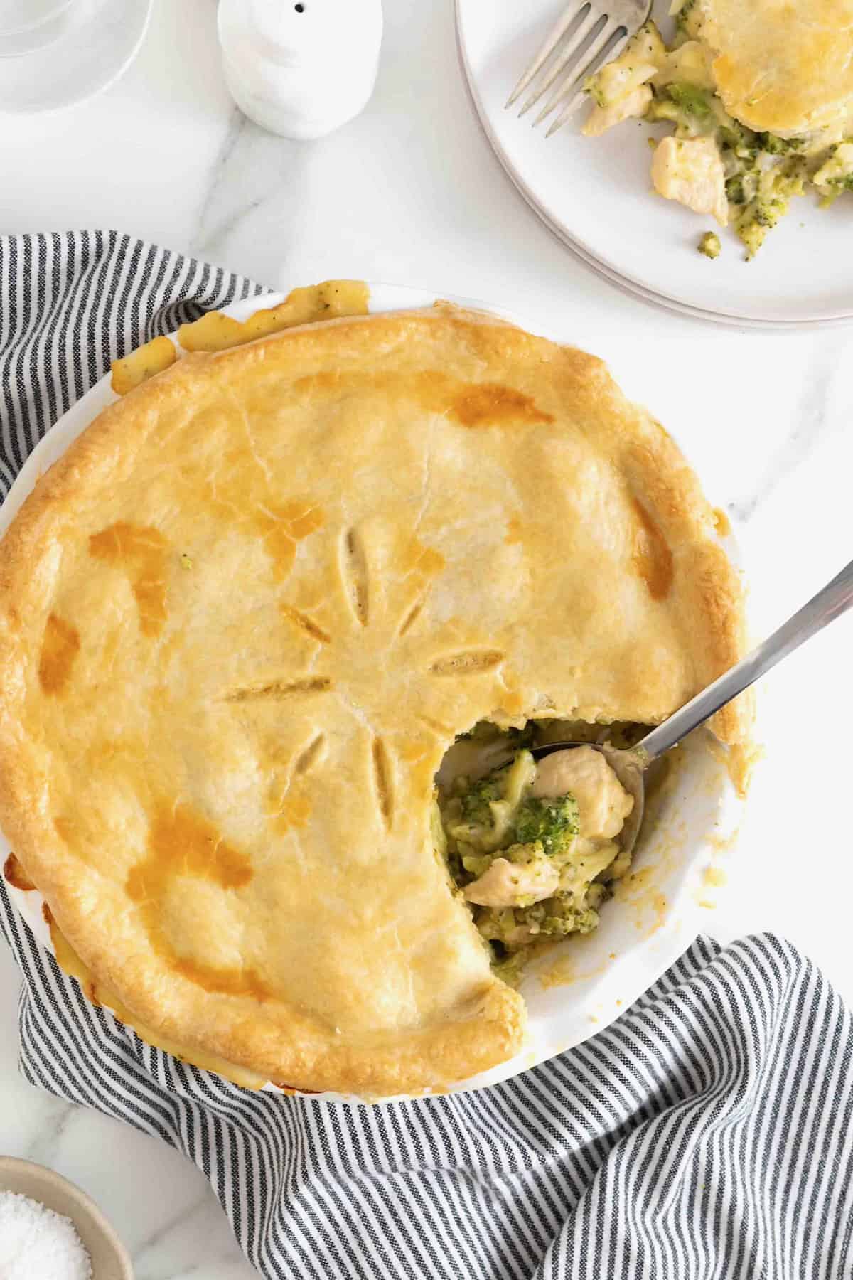 A broccoli and cheese pot pie with a serving taken out of it on a white marble counter.