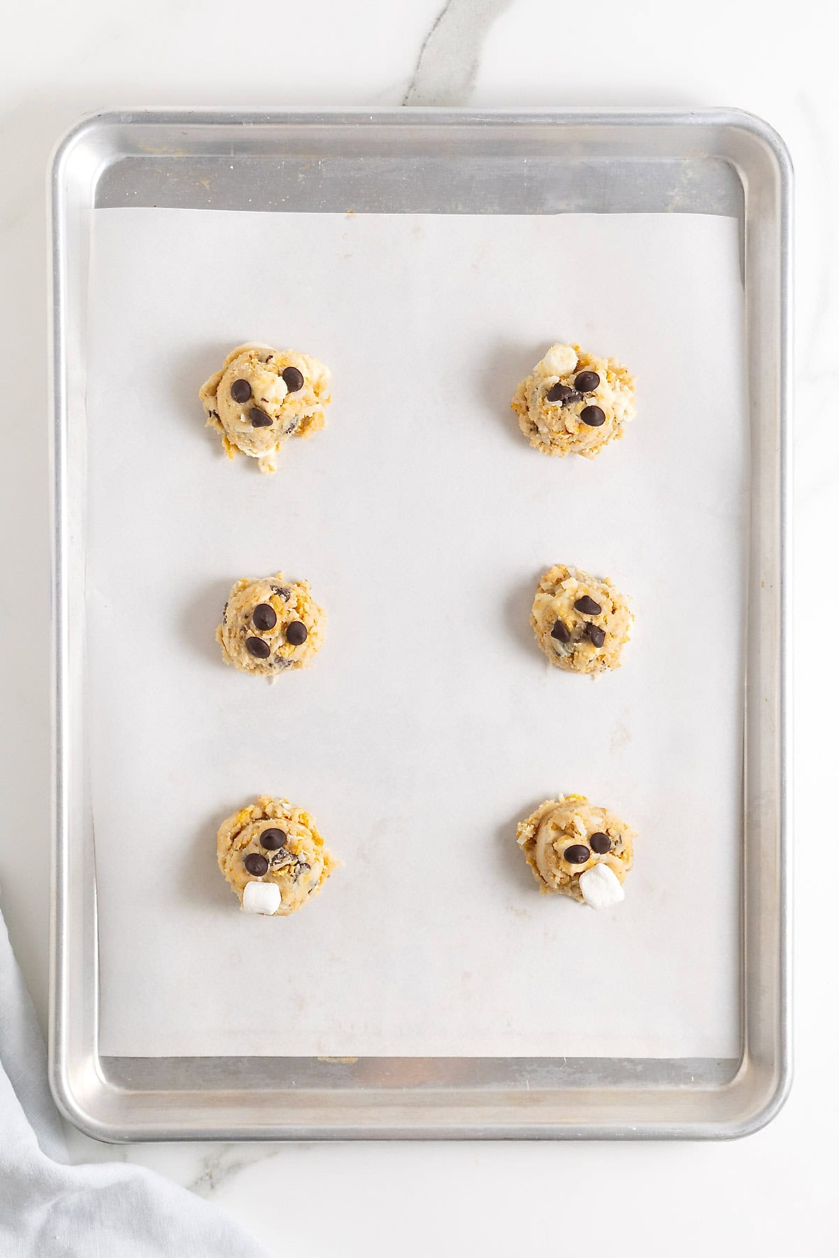Six chocolate chip corn flake coconut cookie dough balls on a parchment lined baking sheet.
