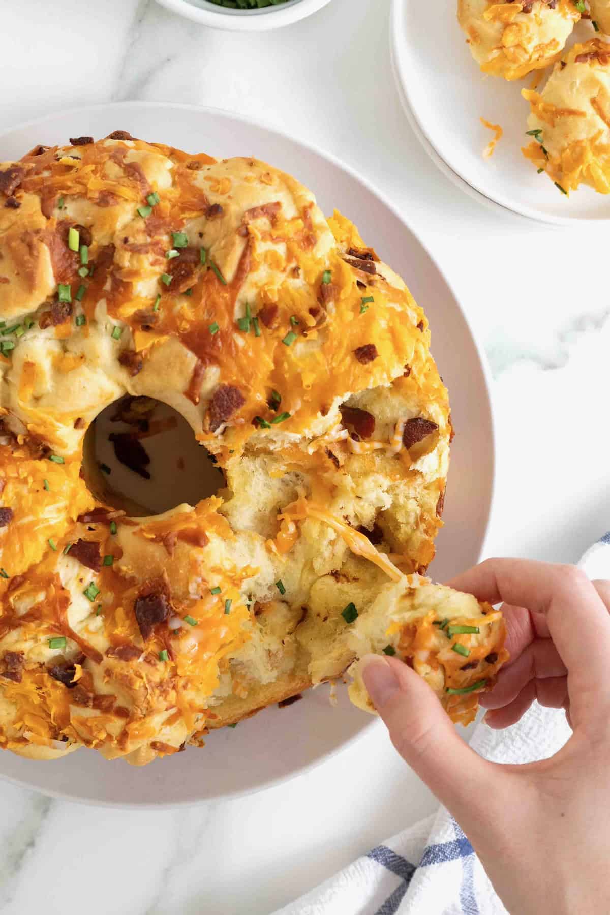 A piece of monkey bread being pulled away from the bacon cheddar monkey bread on a white plate.