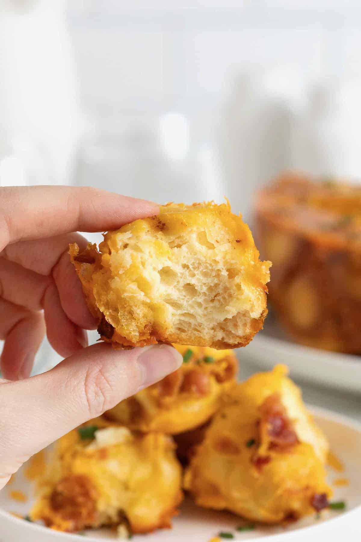 A piece of bacon cheddar monkey bread between a finger and thumb. The bread has a bite out of it revealing a soft inner texture.
