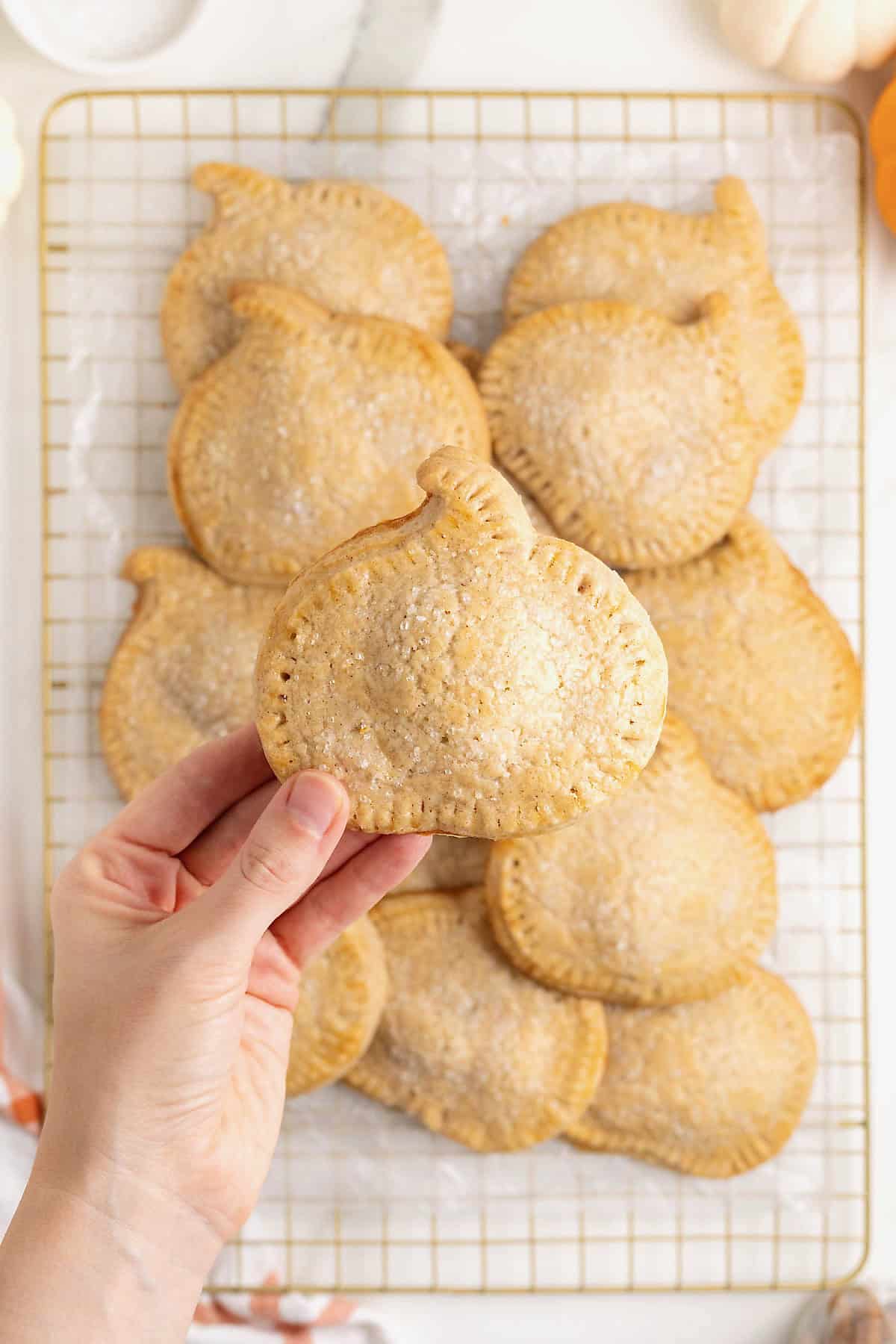A hand holding a pumpkin chocolate hand pie over a pile of pumpkin shaped hand pies on a gold toned cooling rack.