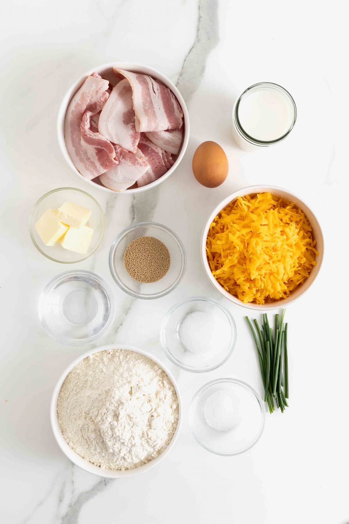 Ingredients to make a bacon cheddar monkey bread in small glass dishes on a white marble counter.