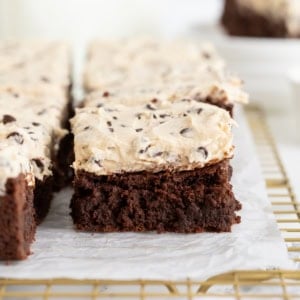 Rich fudge brownies topped with cookie dough frosting on a parchment lined gold-toned cooling rack.