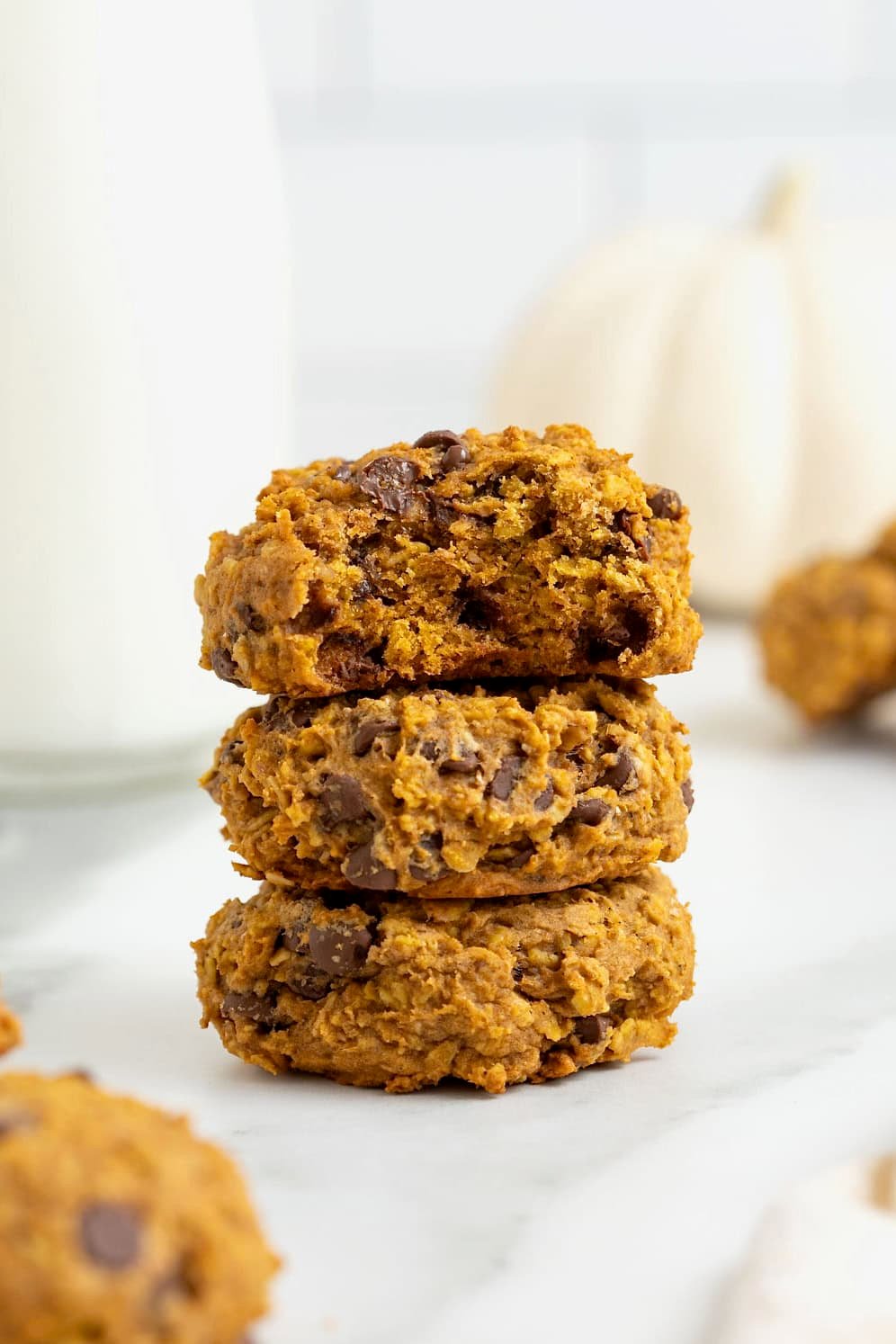 A stack of three pumpkin oatmeal chocolate chip cookies on a white marble counter. The top cookie has a bite out of it.
