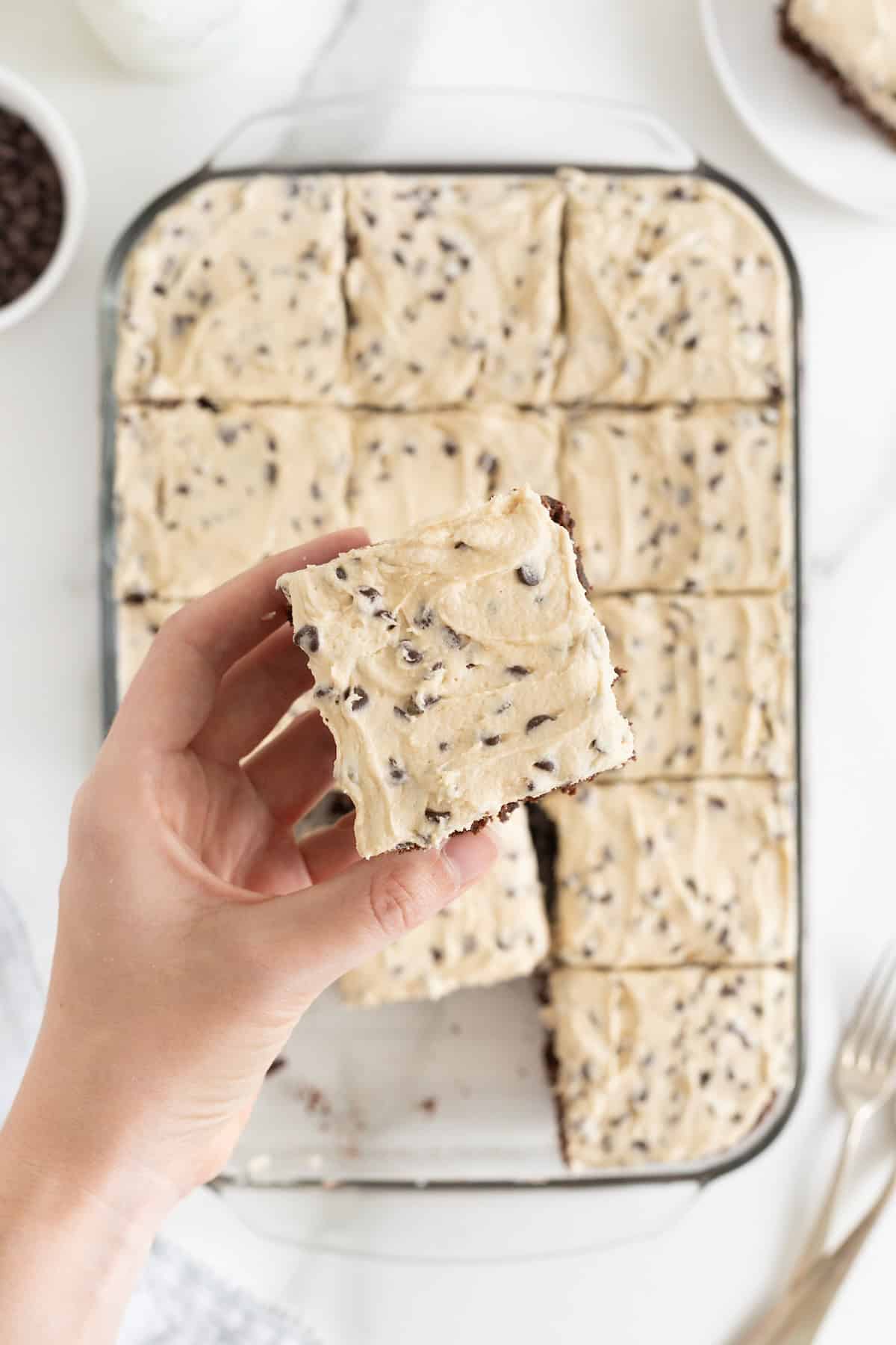 A hand holding a cookie dough frosted brownie over a glass baking dish of brownies.