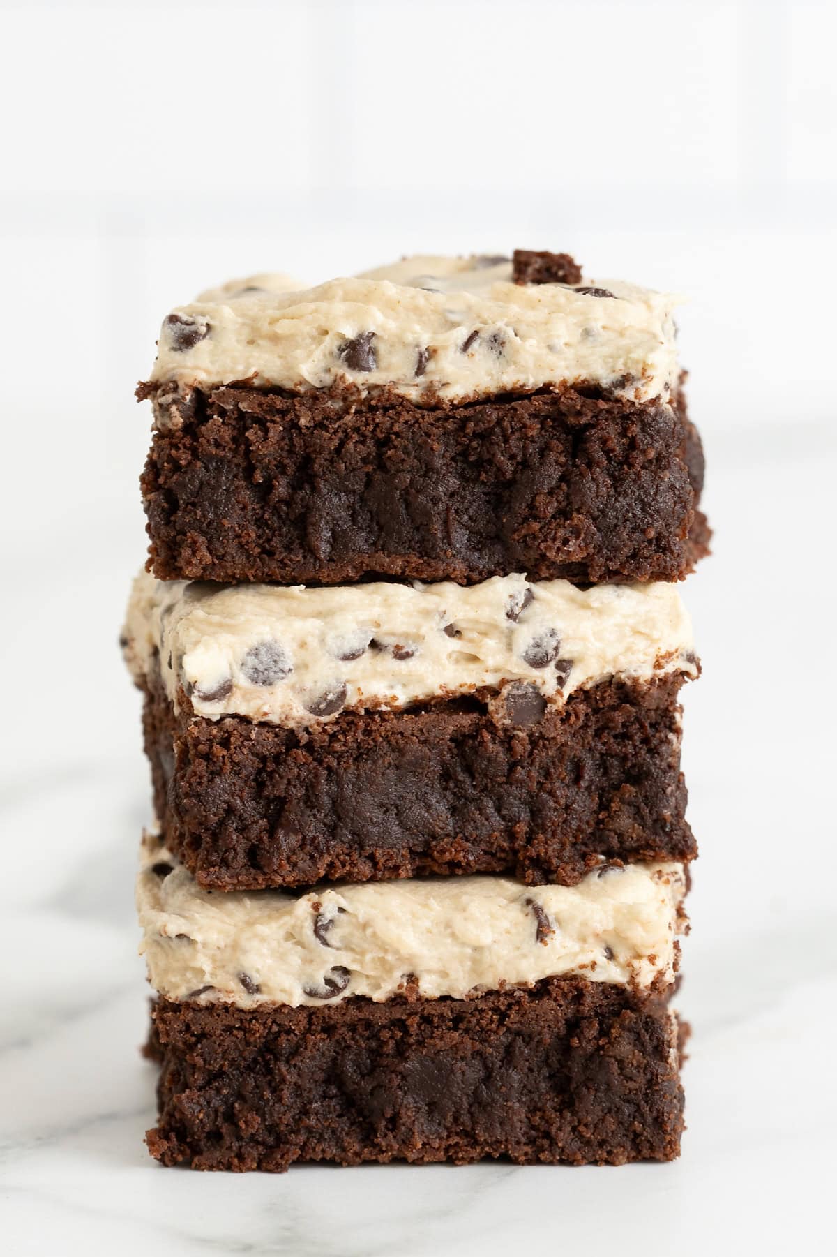 Three chocolate chip cookie dough frosted fudge brownies stacked a white marble counter.