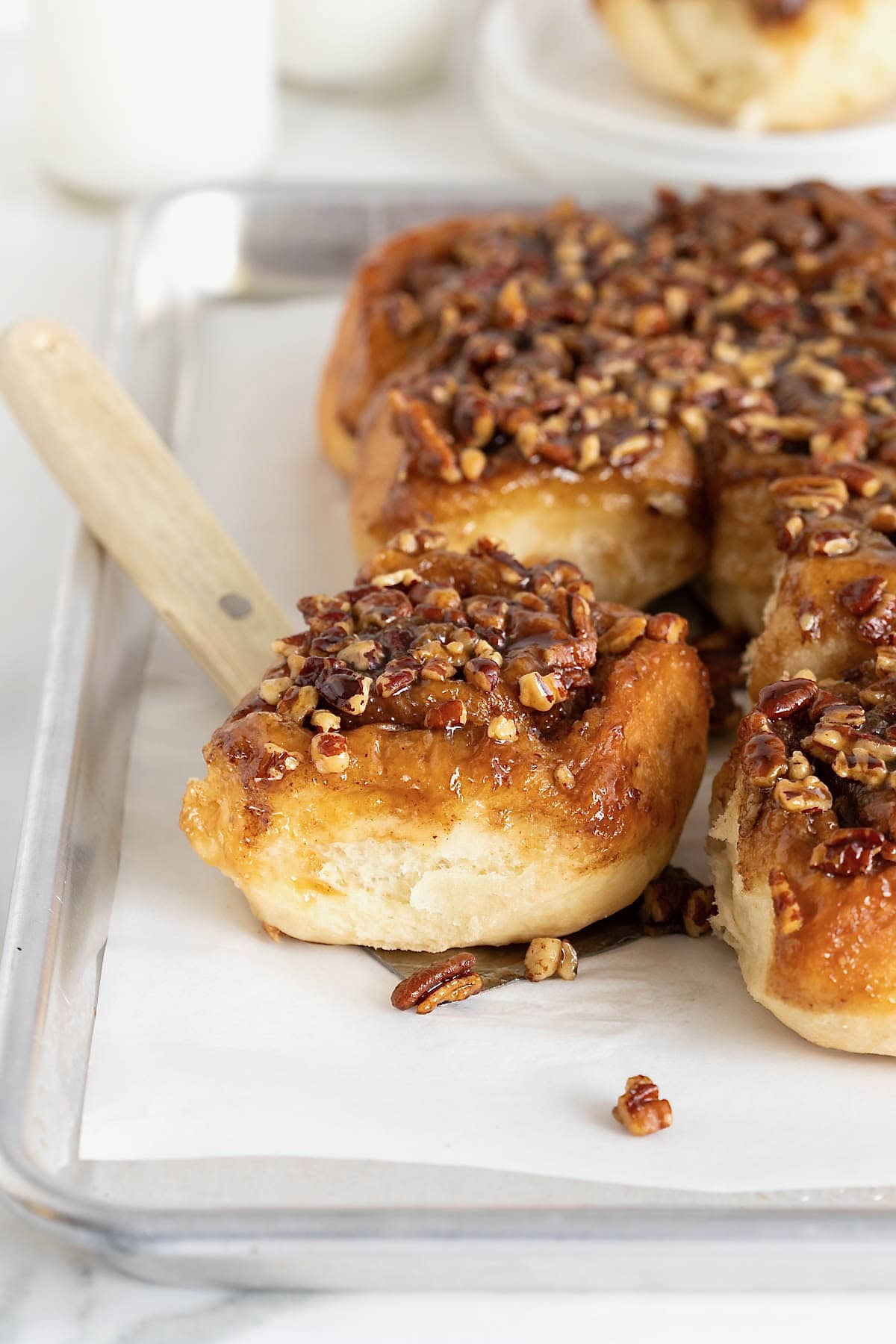 A sticky bun on a spatula resting on a parchment lined aluminum baking sheet of sticky buns.
