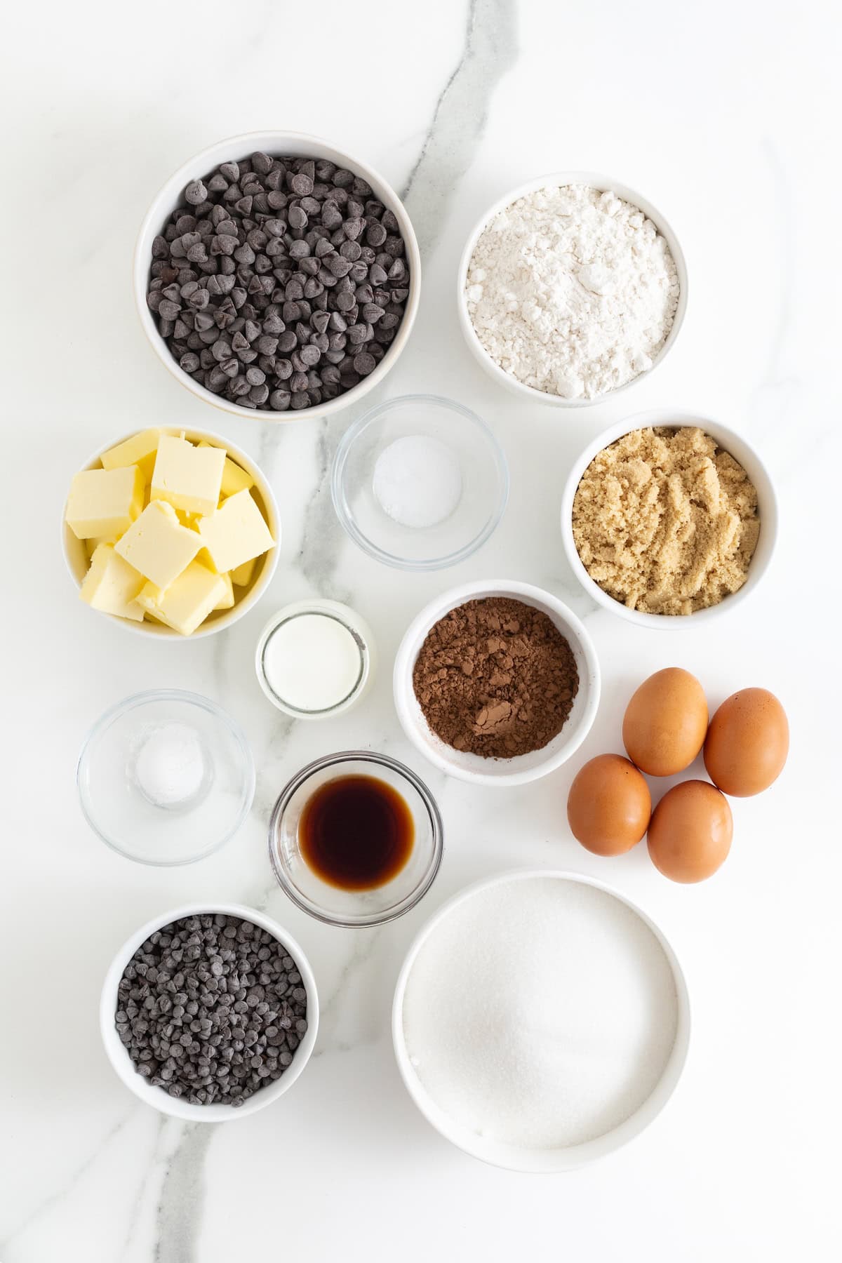 Ingredients to make cookie dough frosted fudge brownies in small white and clear glass dishes on a white marble counter.
