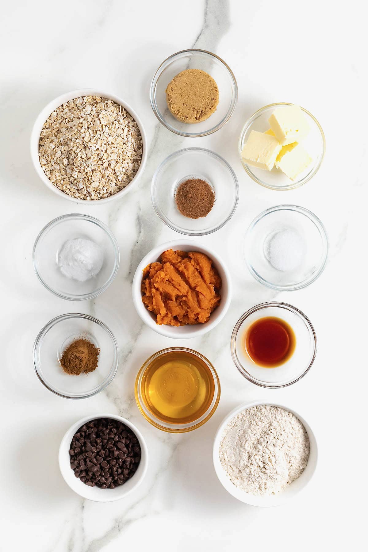 Ingredients to make pumpkin oatmeal chocolate chip cookies in small glass dishes on a white marble counter.