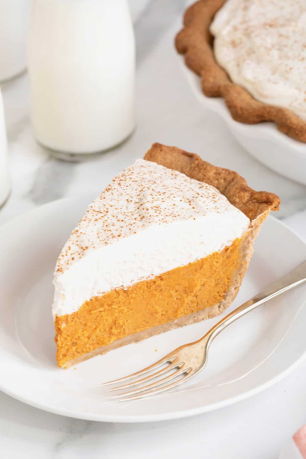 A slice of pumpkin pie with a layer of whipped topping on a white dessert plate. There is a fork next to the pie.