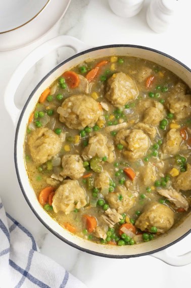 A white Dutch oven of turkey and corn bread dumplings with peas and carrots.