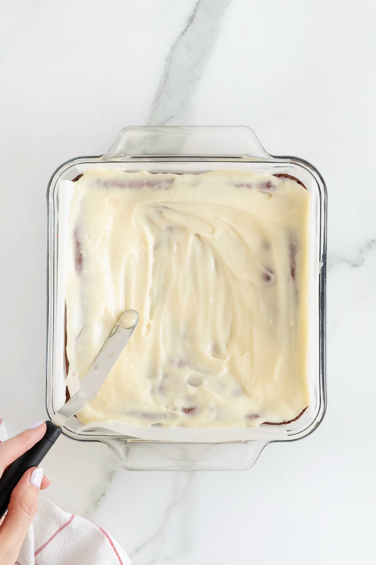 Cream cheese frosting being spread over red velvet brownies in a square glass baking dish.