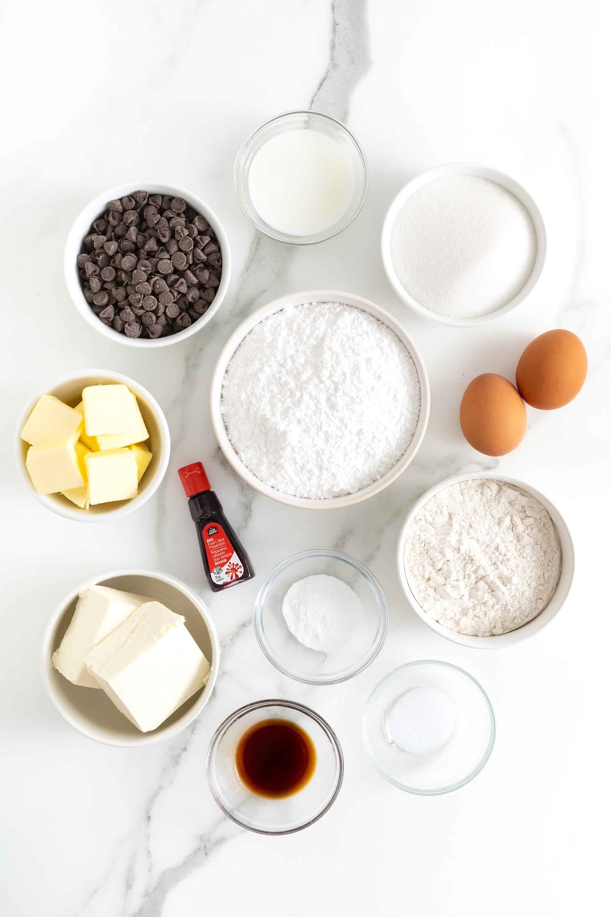 Ingredients to make red velvet brownies with cream cheese frosting in small white dishes on a white marble counter.