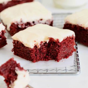 Frosted red velvet brownies on a parchment lined cooling rack. one of the brownies has a bite out of it.