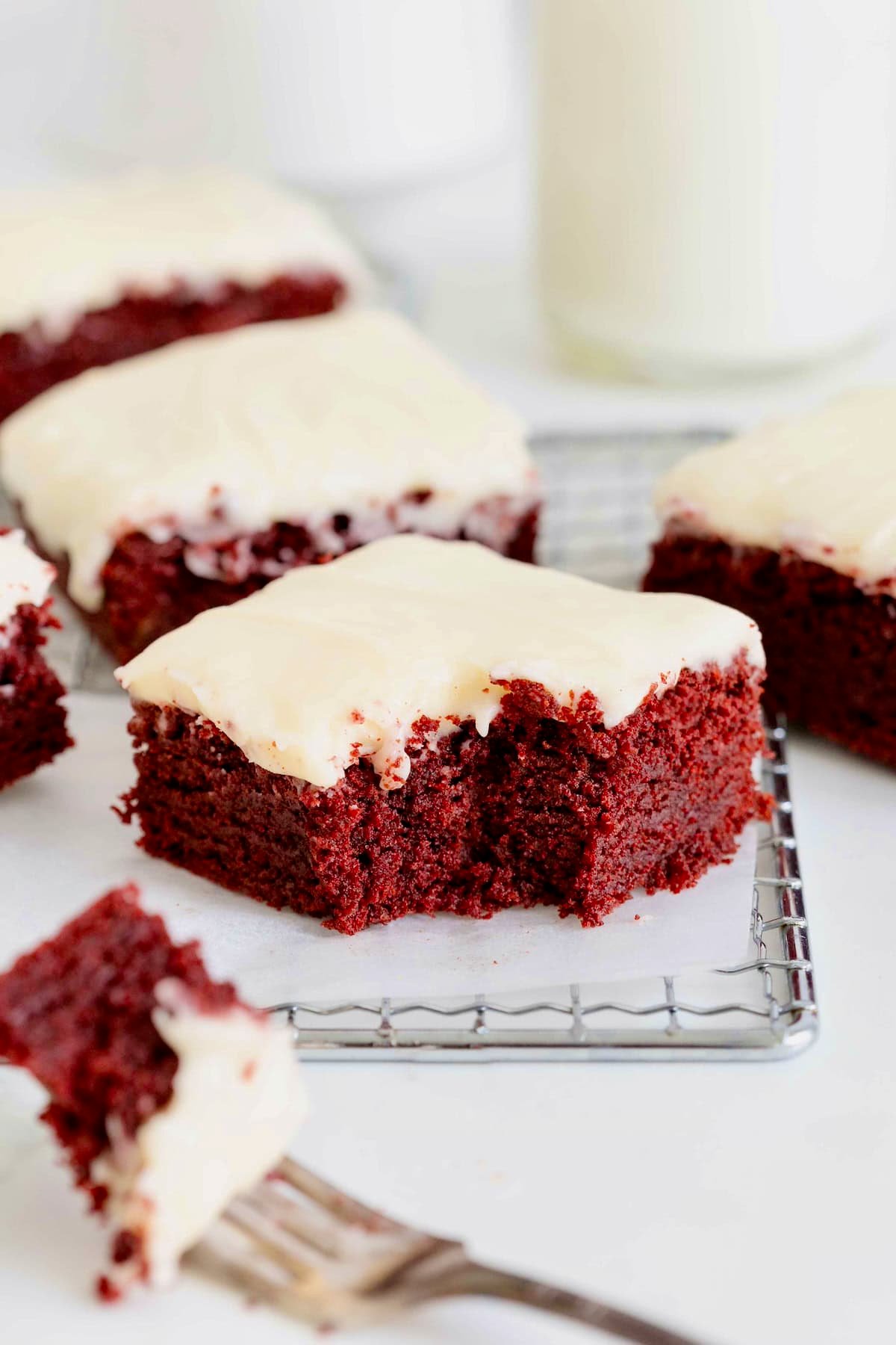 Frosted red velvet brownies on a parchment lined cooling rack. one of the brownies has a bite out of it.