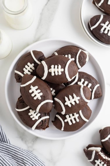 A large white platter of chocolate whoopie pies decorated like footballs.