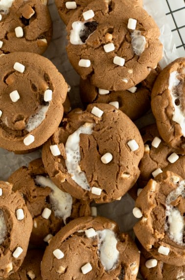 A pile of hot cocoa cookies with mini marshmallows on top on a parchment lined metal cooling rack.
