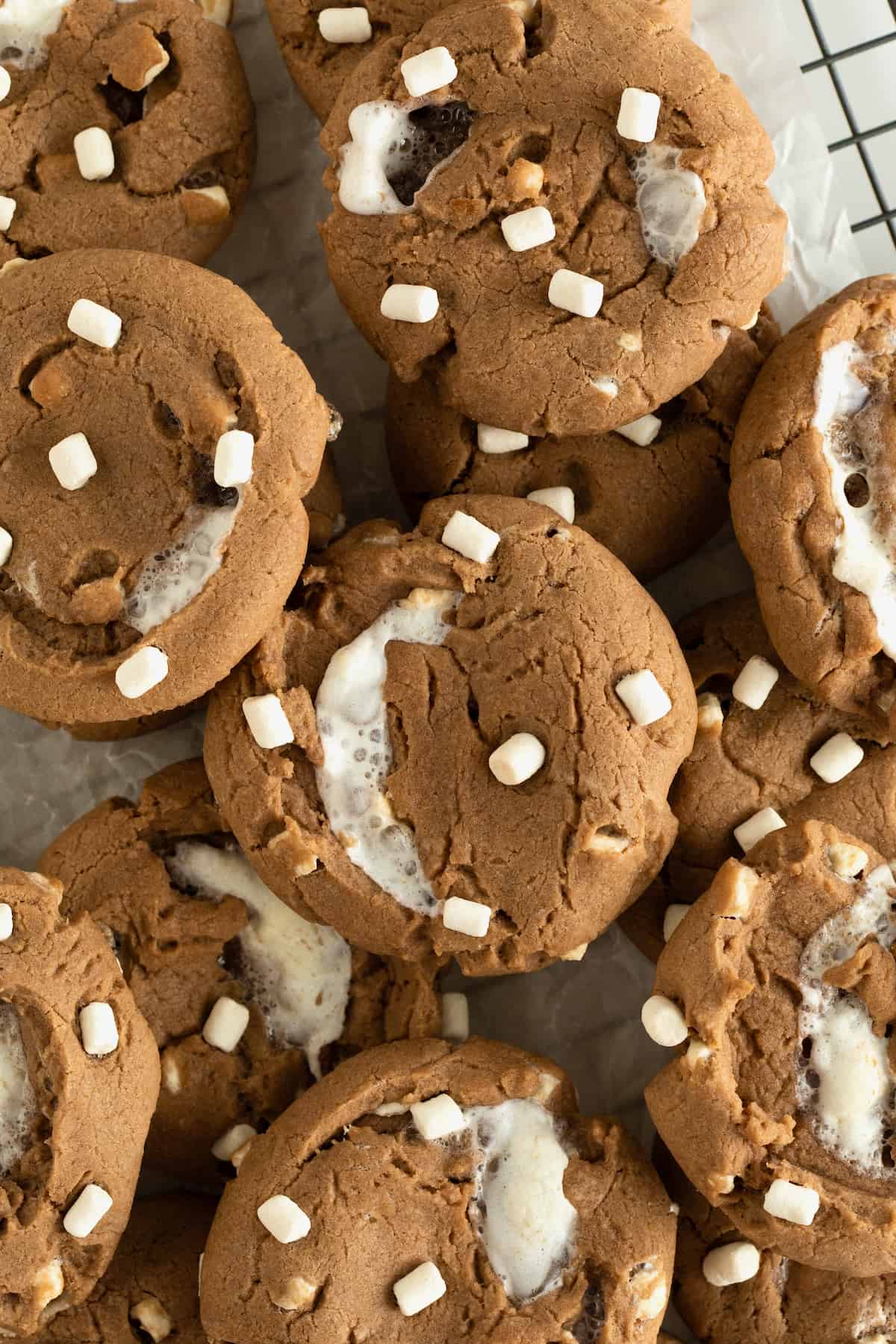 A pile of hot cocoa cookies with mini marshmallows on top on a parchment lined metal cooling rack.