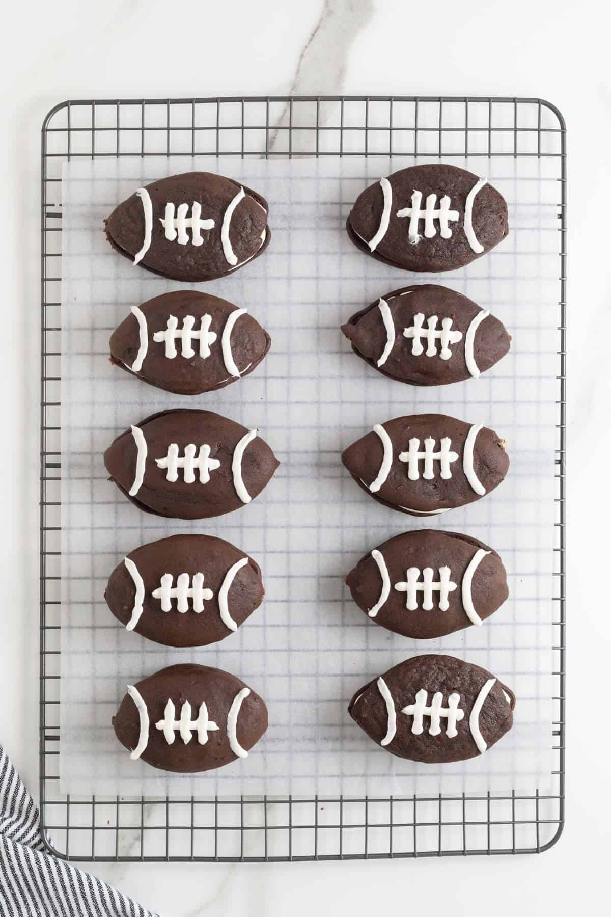 10 football shaped chocolate whoopie pies on a parchment lined cooling rack.