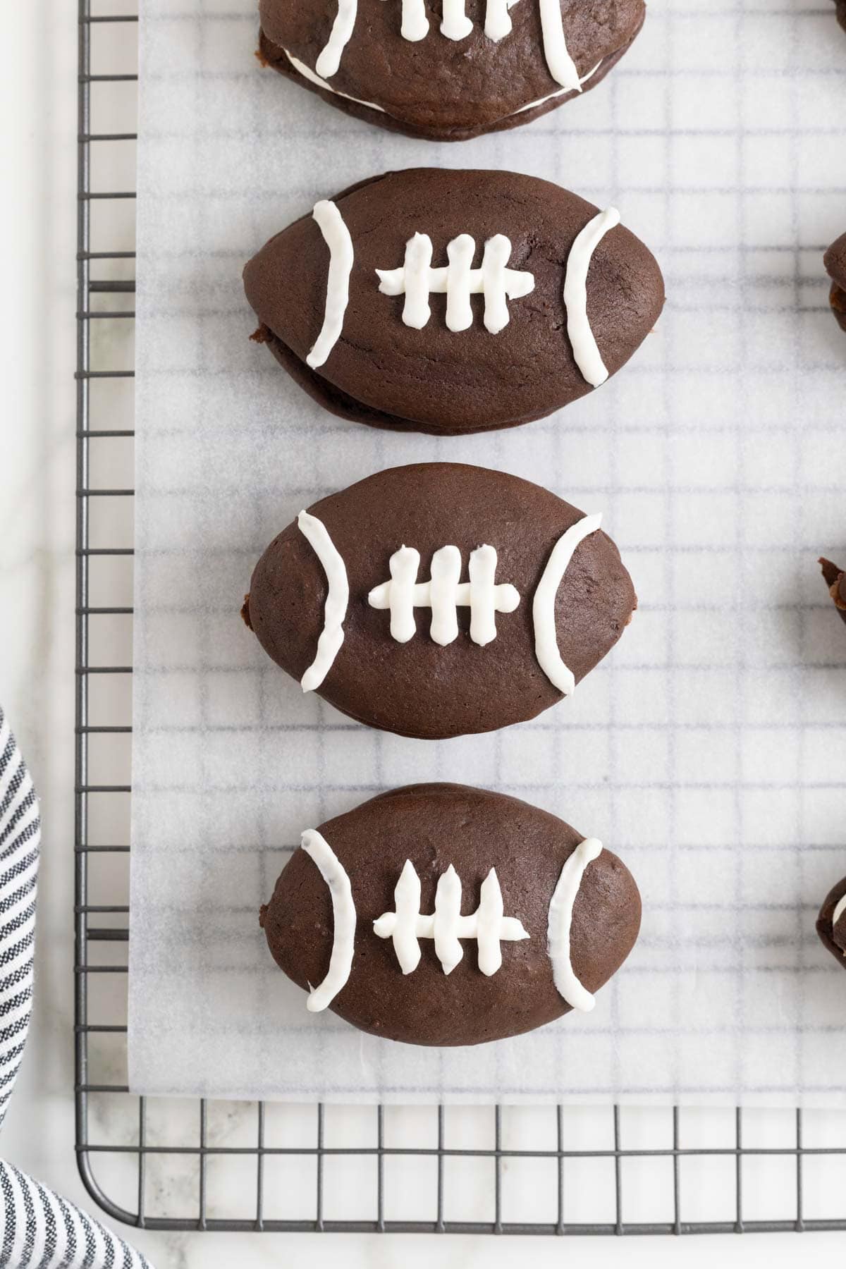 Four football shaped chocolate whoopie pies on a parchment lined cooling rack.