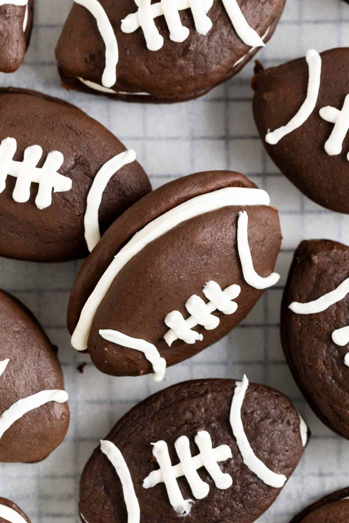 A pile of football shaped chocolate whoopie pies on a parchment lined wire cooling rack.