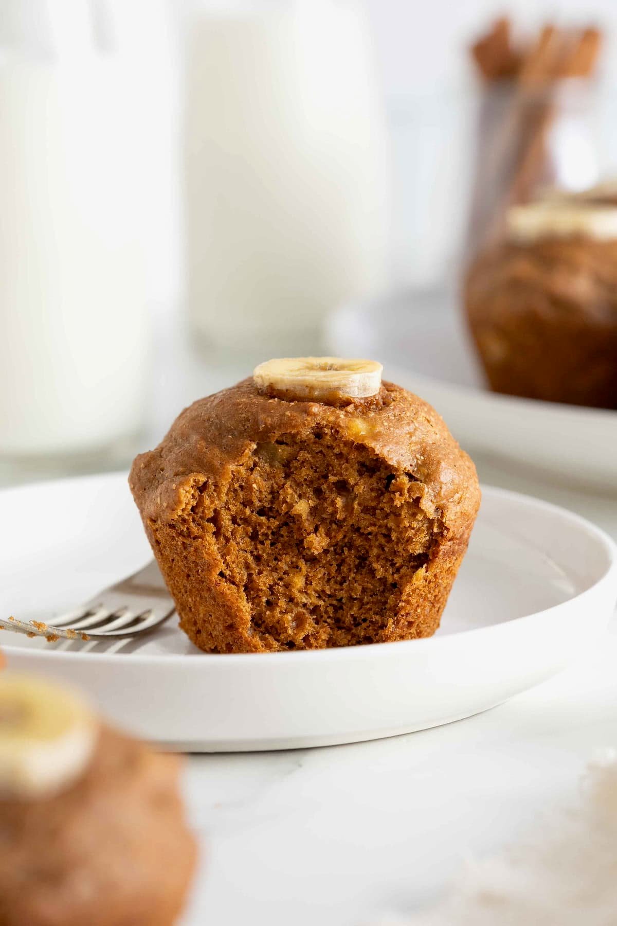A white rimmed plate with a single gingerbread muffin with a bite taken out of it. There is a metal fork next to the muffin on the plate.