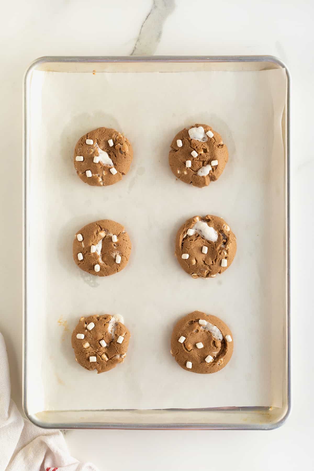 Six hot cocoa cookies on a parchment lined aluminum baking sheet.