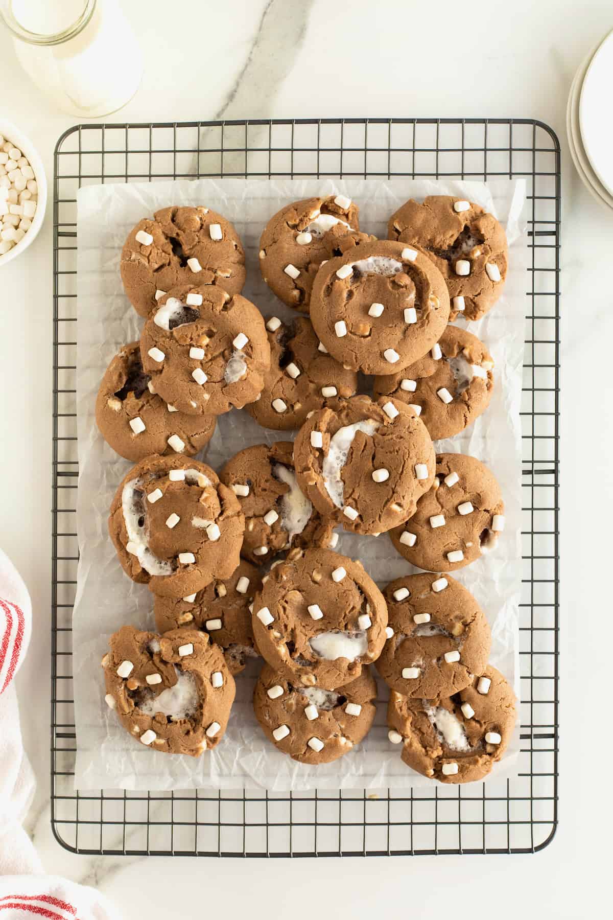 A pile of hot cocoa cookies with mini marshmallows on top on a parchment lined metal cooling rack.
