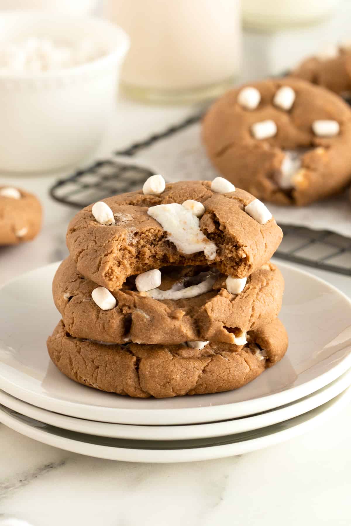 A stack of three hot cocoa cookies on a white plate. The top cookie has a bite out of it revealing melting marshmallow.