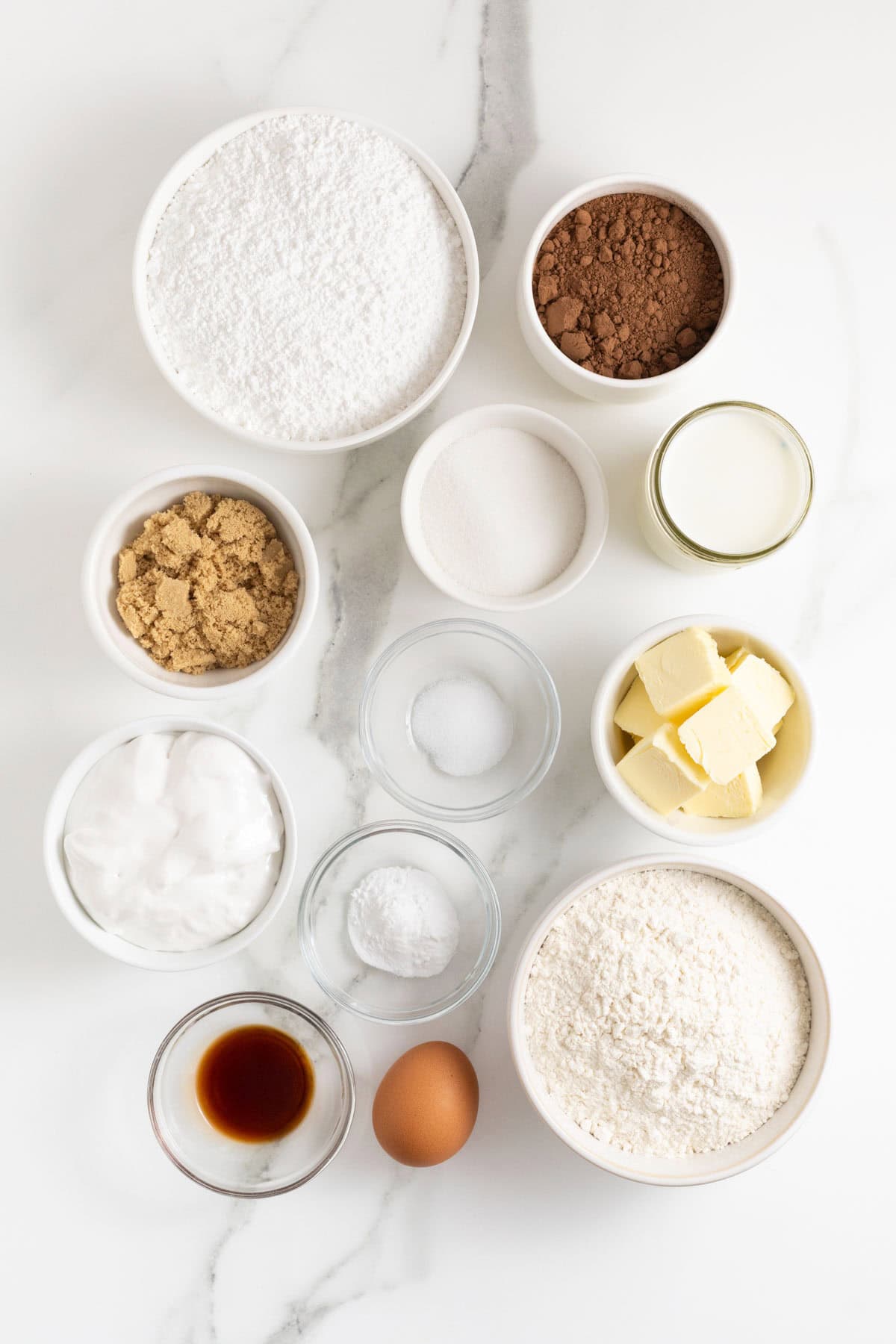 Ingredients to make chocolate football shaped whoopie pies in small white dishes on a white marble counter.