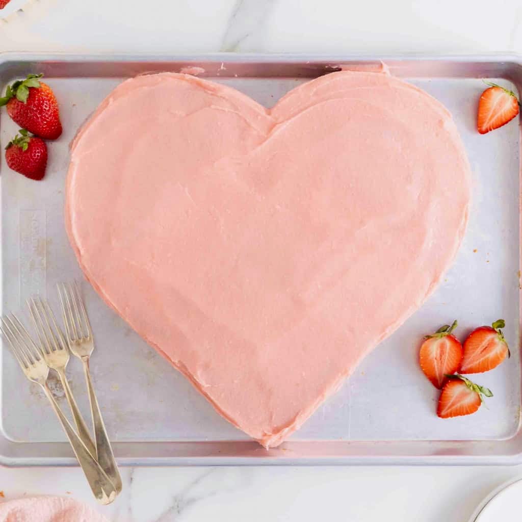 A heart shaped cake with pink icing on a baking sheet.