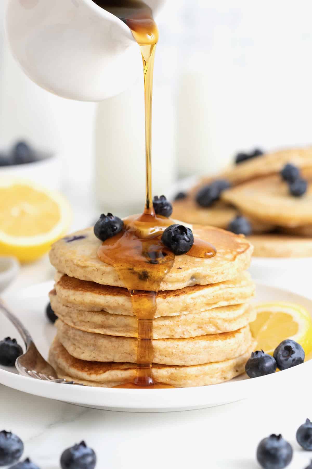 Syrup being poured over a stack of four blueberry lemon ricotta pancakes.