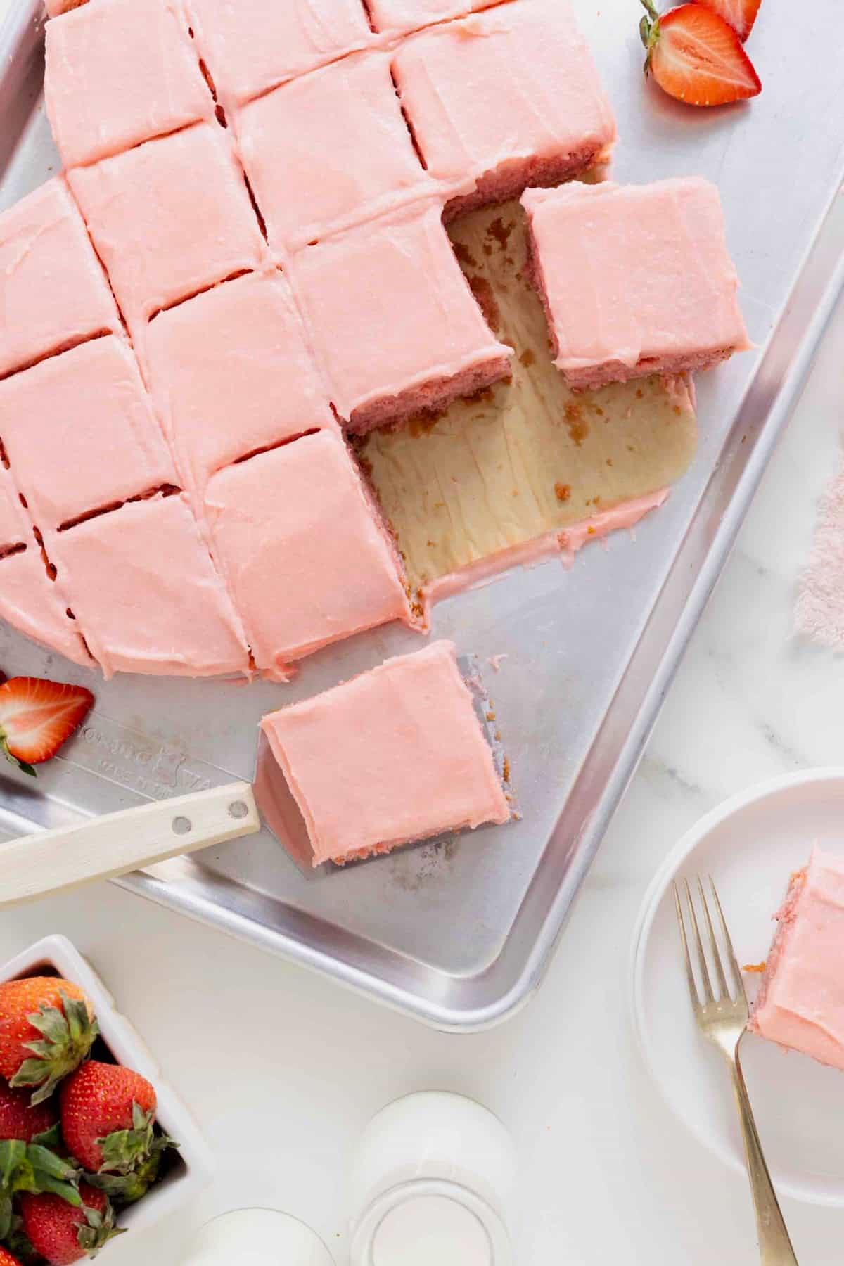 A heart shaped cake with light pink frosting cut into squares. A spatula is lifting one slice from the cake.