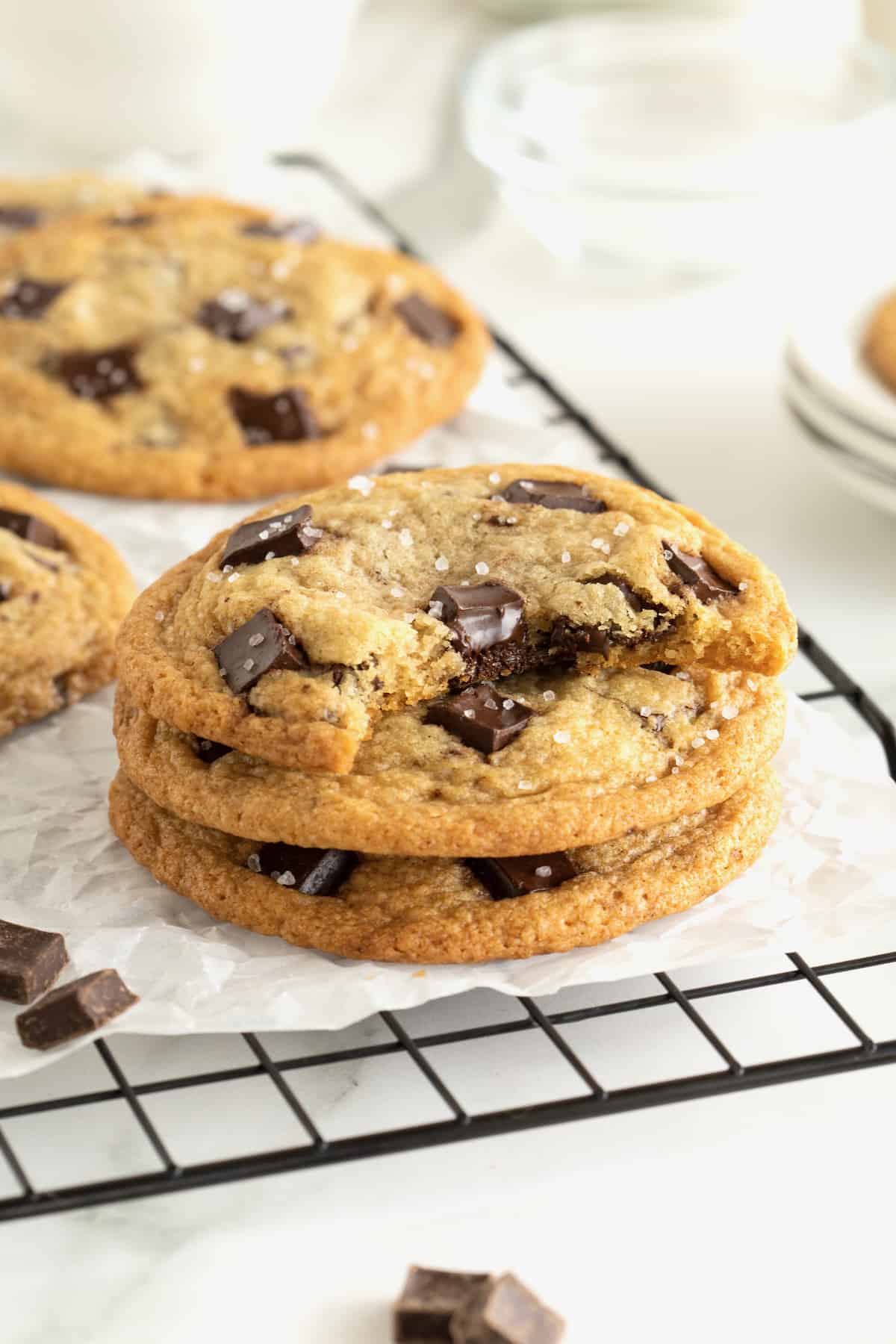 Three chocolate chunk cookies in a stack on a parchment lined cooling rack. The top cookie has a bite out of it.