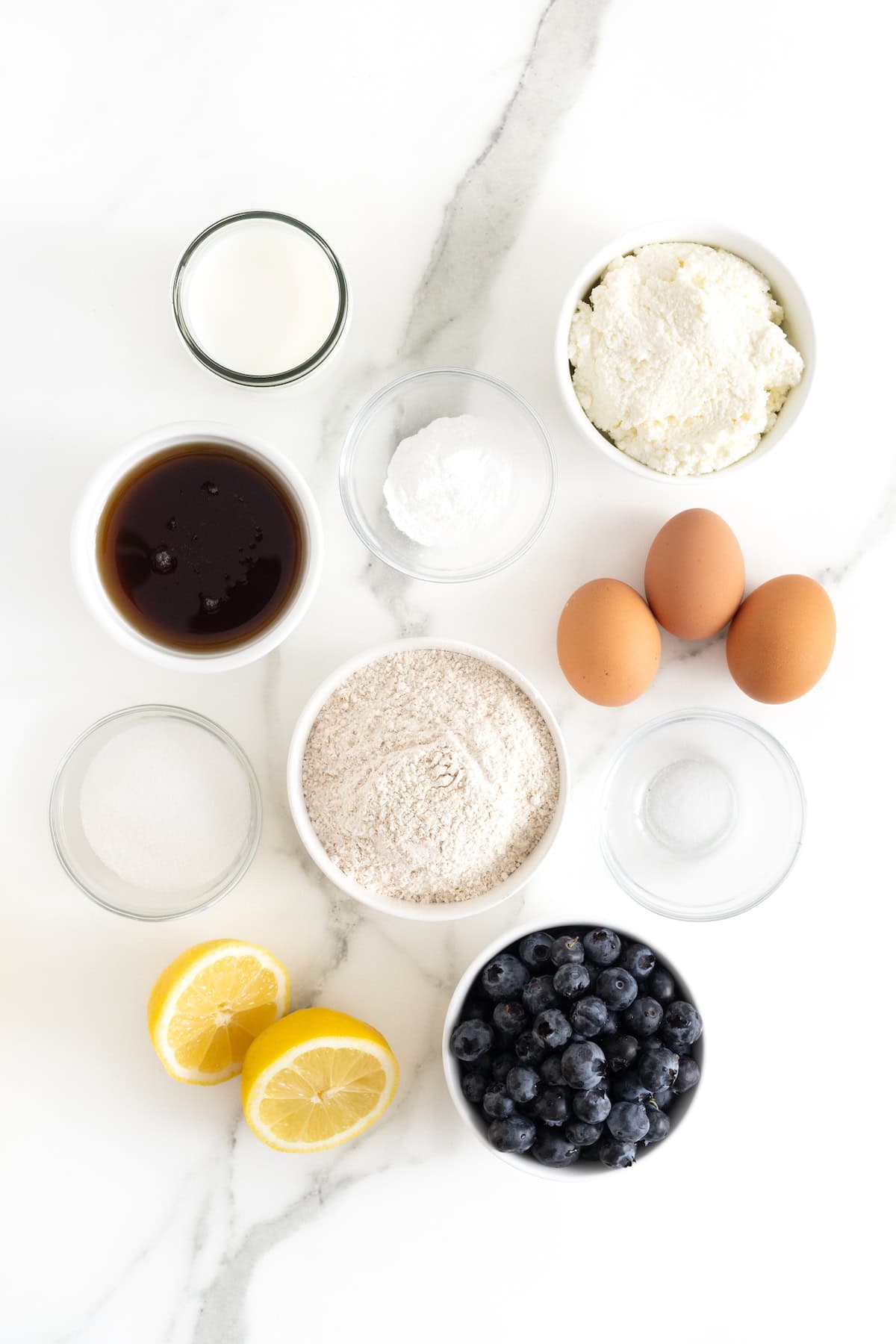 Ingredients to make whole wheat blueberry lemon ricotta pancakes in small glass dishes on a white marble counter.