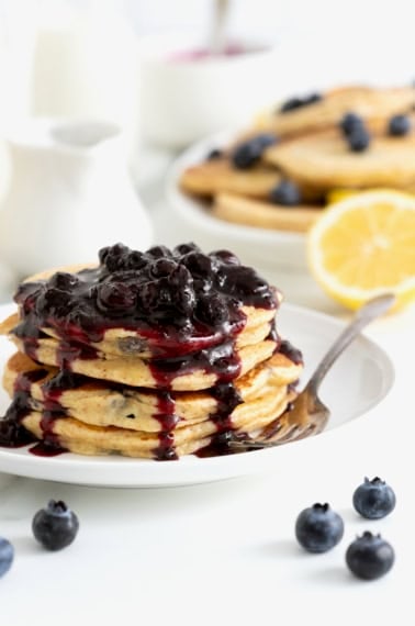 Blueberry pancakes stacked on a white plate covered in blueberry syrup.