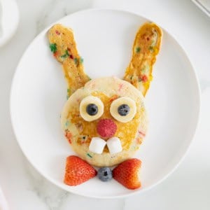 A white plate with pancakes made to look like the Easter bunny, with a strawberry bowtie.