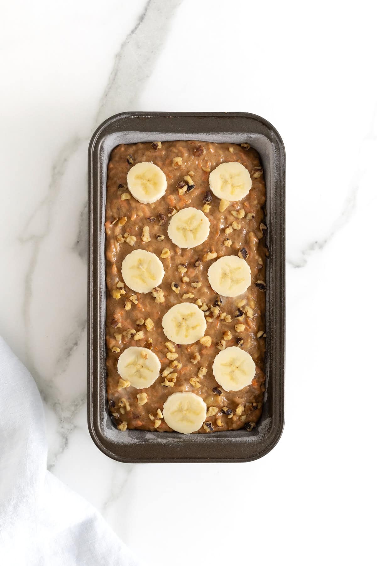 A dark loaf pan filled with banana nut carrot cake bread batter garnished with banana slices.