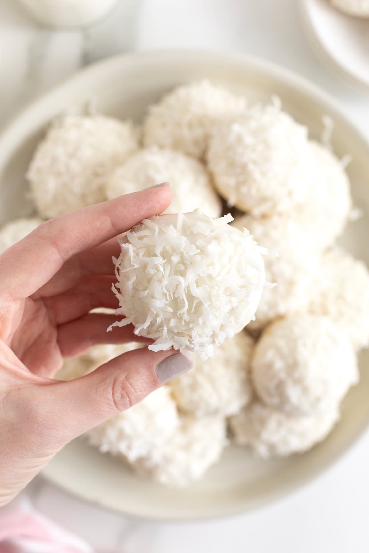 A hand holding a white chocolate dipped coconut cookie over a white rimmed plate of cookies.