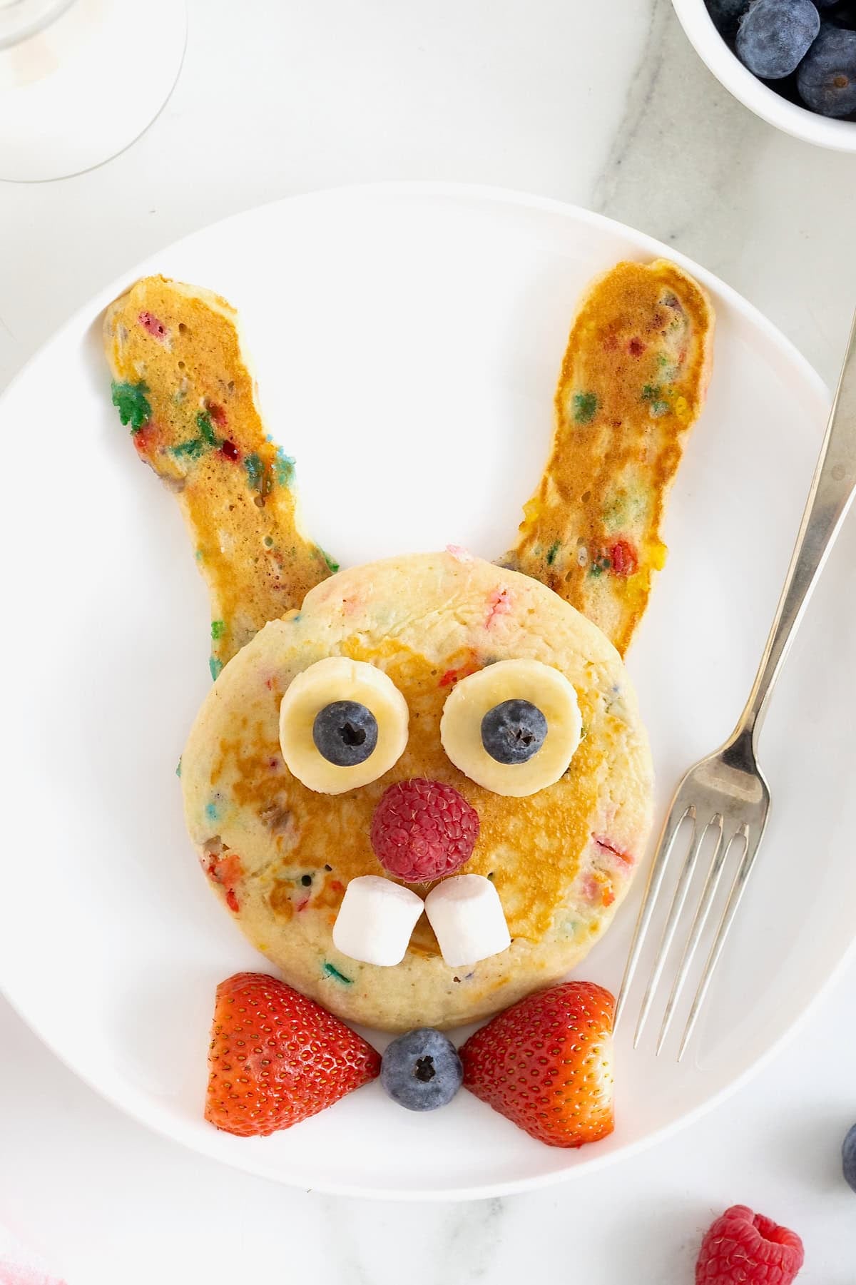 A white plate with pancakes made to look like the Easter bunny, with a strawberry bowtie. There is a metal fork next to the pancakes.