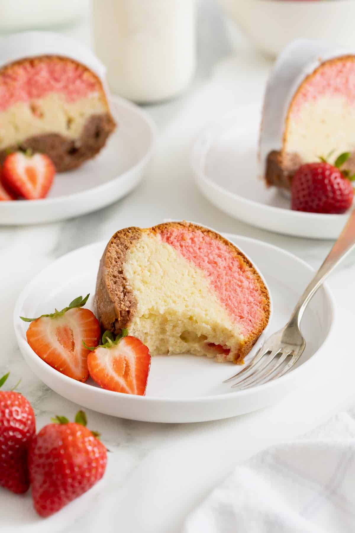 A slice of Neapolitan pound cake on a white dessert plate with a fork and a halved strawberry.