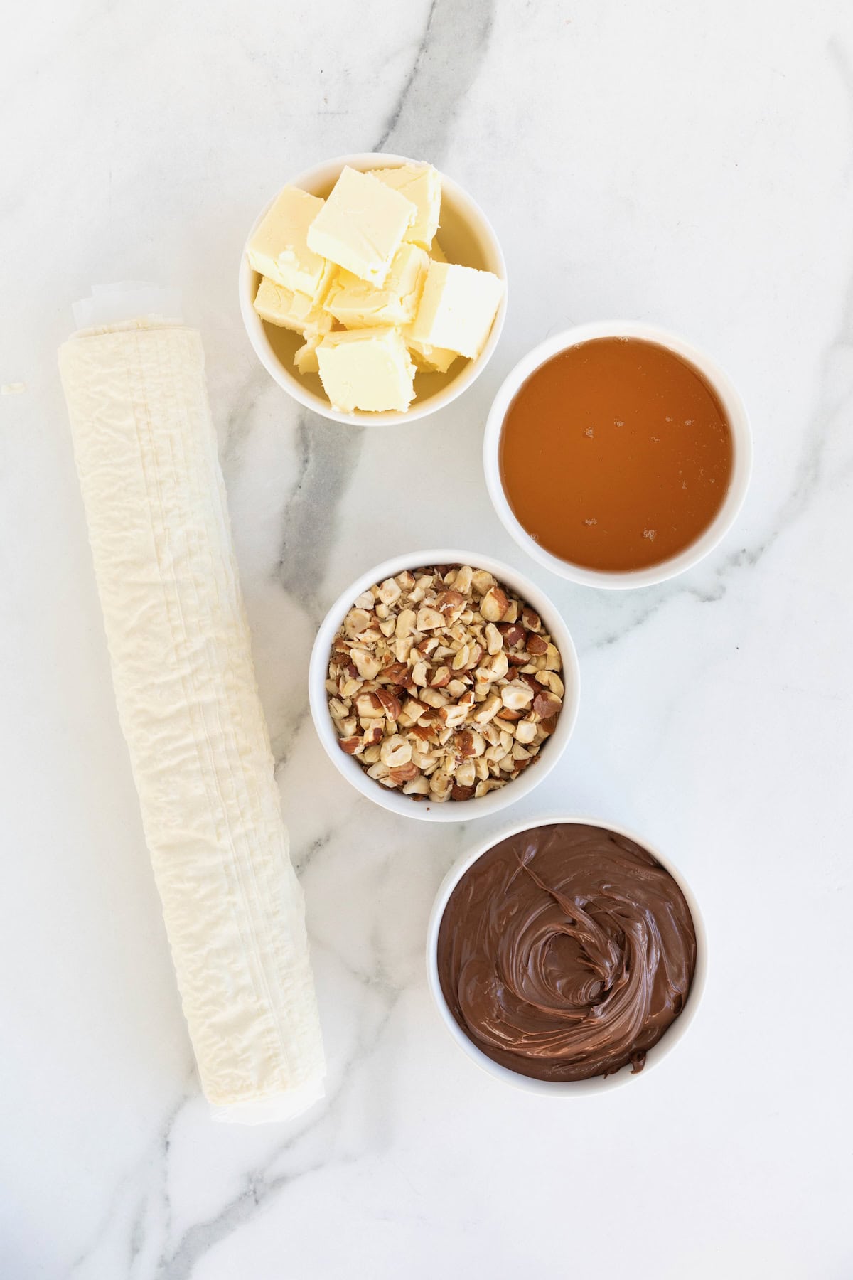 Ingredients to make Nutella baklava in small white dishes on a white marble counter.