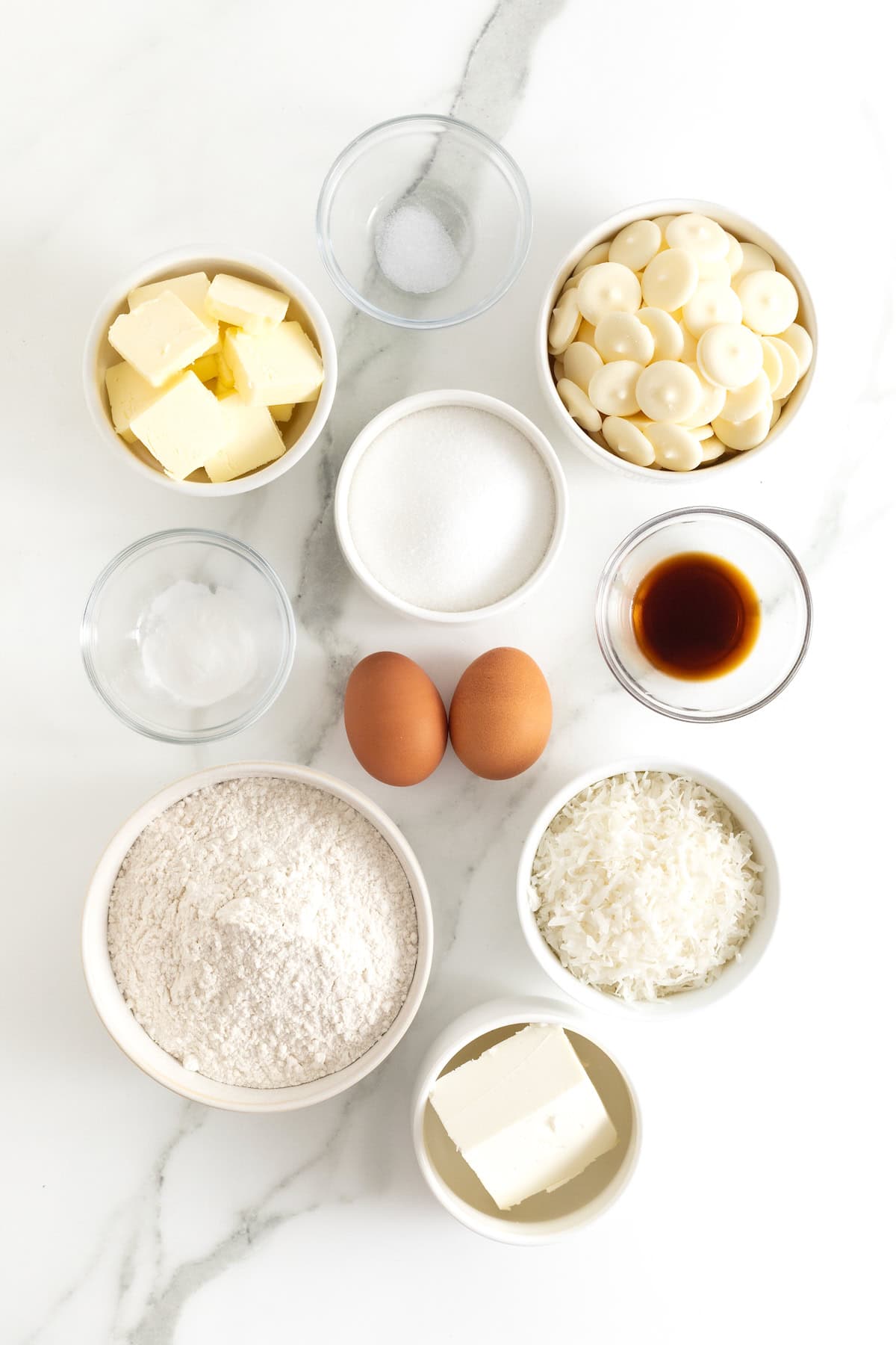 Ingredients to make white chocolate dipped coconut cookies in small glass dishes on a white marble counter.
