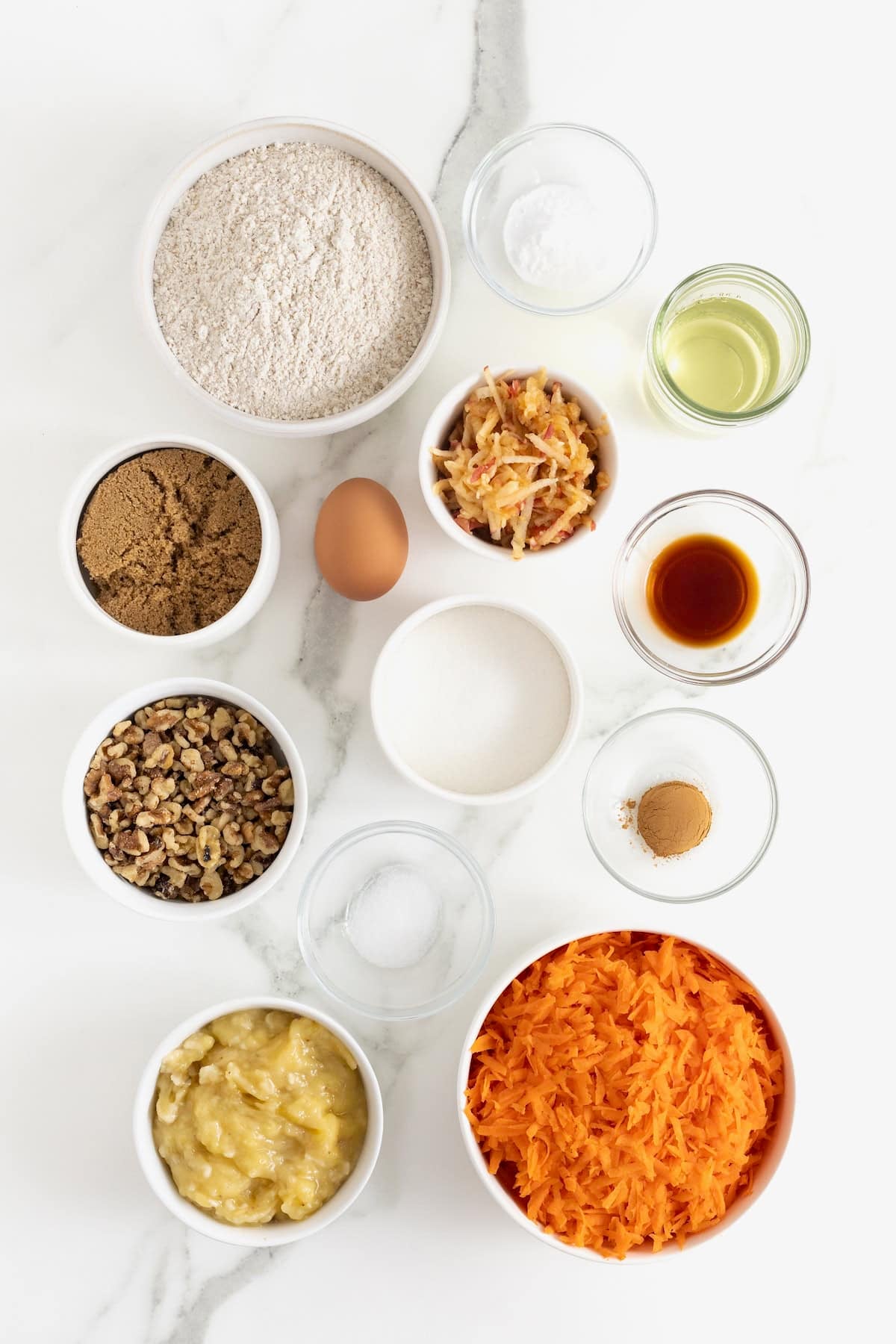Ingredients to make whole wheat banana nut carrot cake bread in small glass dishes on a white marble counter.