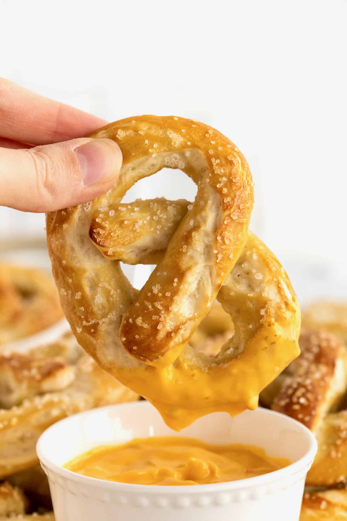 A hand holding a pretzel that has just been dipped in cheese sauce. A plate full of soft pretzels with a small dish of cheese sauce is in the background.