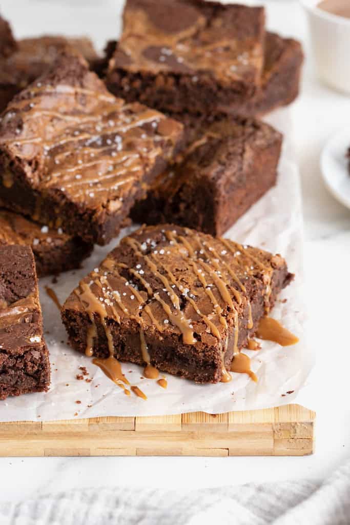 A pile of salted dulce de leche brownies drizzled with caramel on a parchment lined wooden cutting board.
