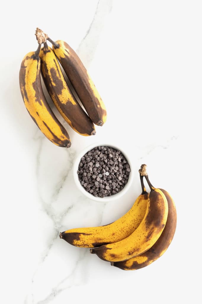 Two bunches of overripe bananas on either side of a small white dish of mini chocolate chips.
