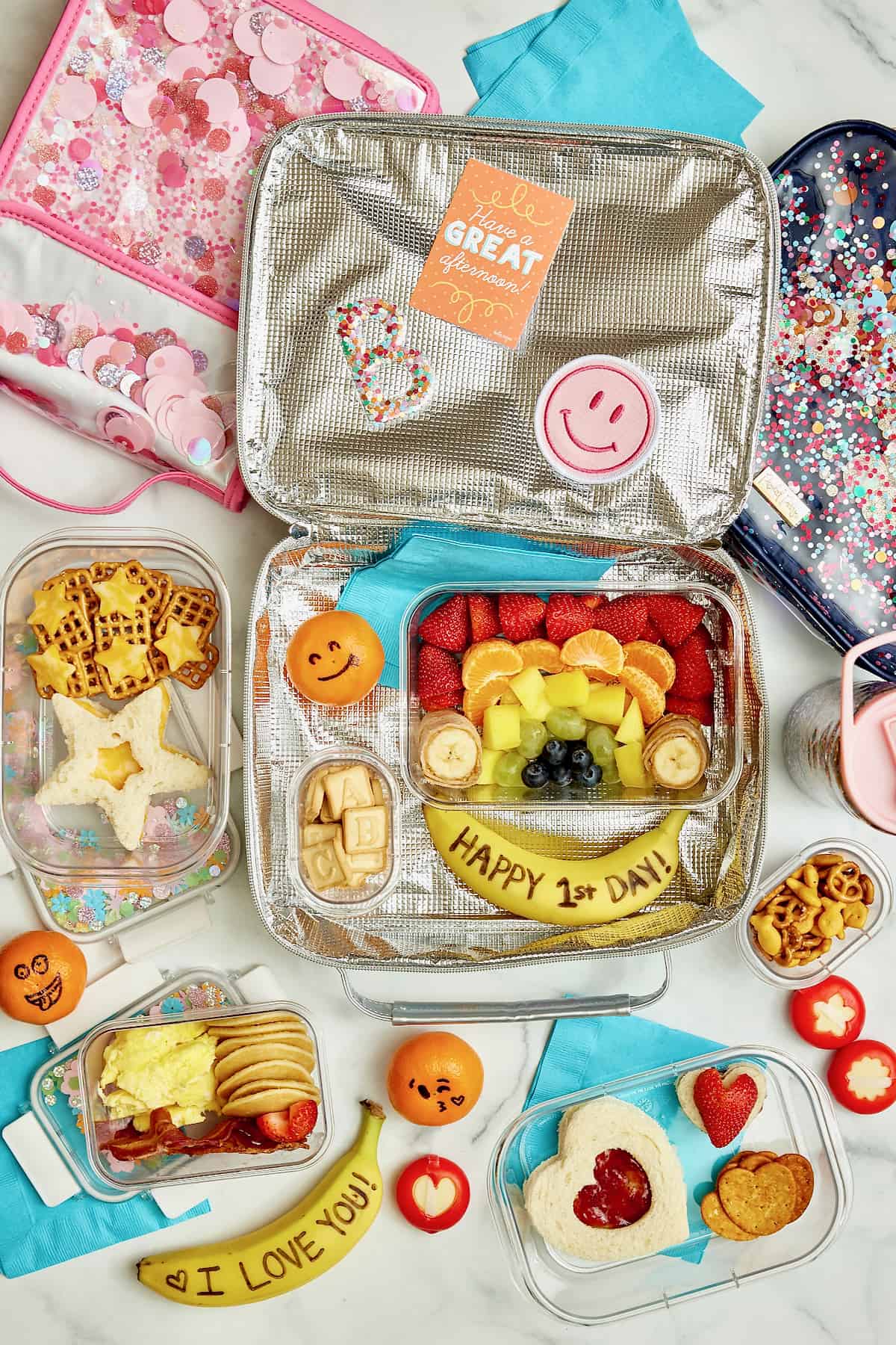 A lunch box with fruit arranged in a rainbow and a banana that says Happy First Day of School. Mini pancakes, scrambled eggs and sliced strawberries in a lunch box. A star shaped cheese sandwich, star shaped cheese slices on pretzels.