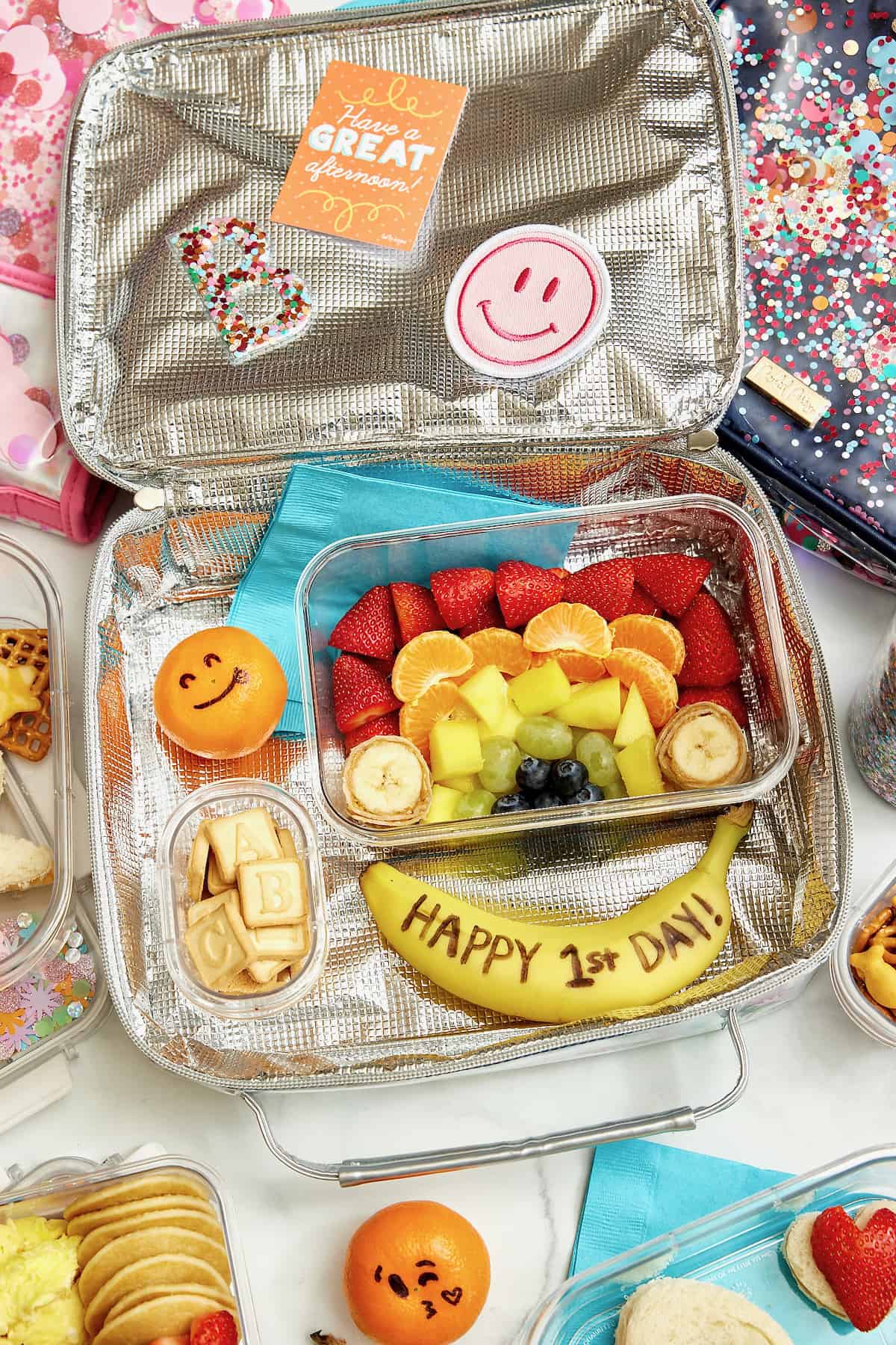 A lunch box with fruit arranged in a rainbow and a banana that says Happy First Day of School